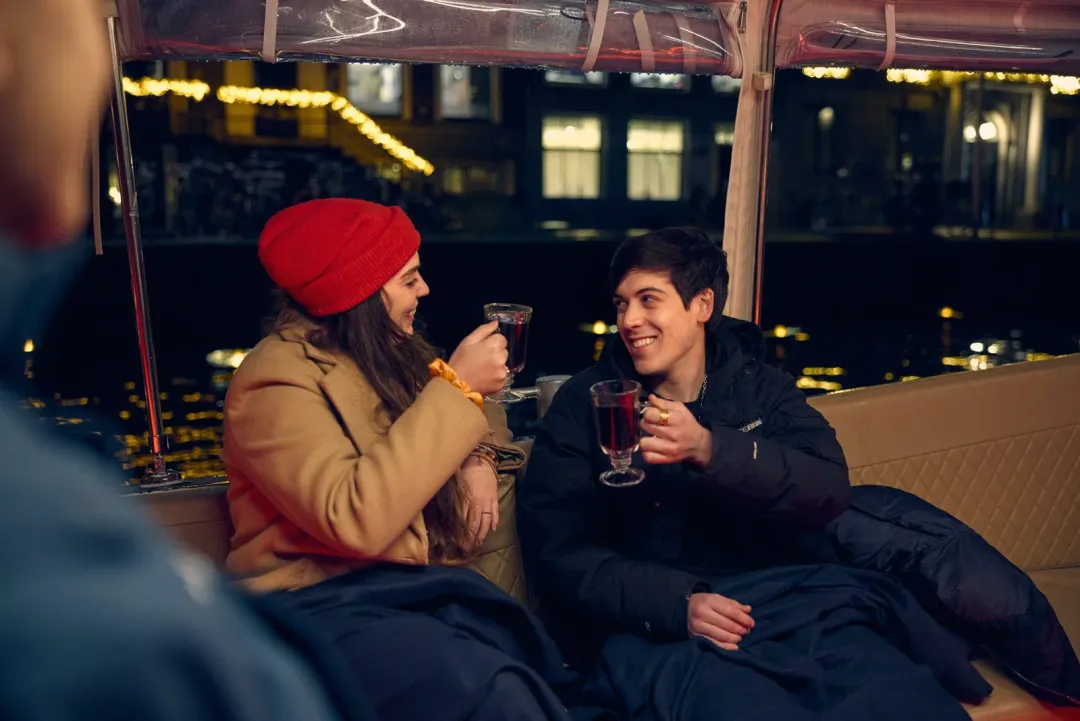 Two guests in winter coats hold hot drinks under blankets on a boat at night