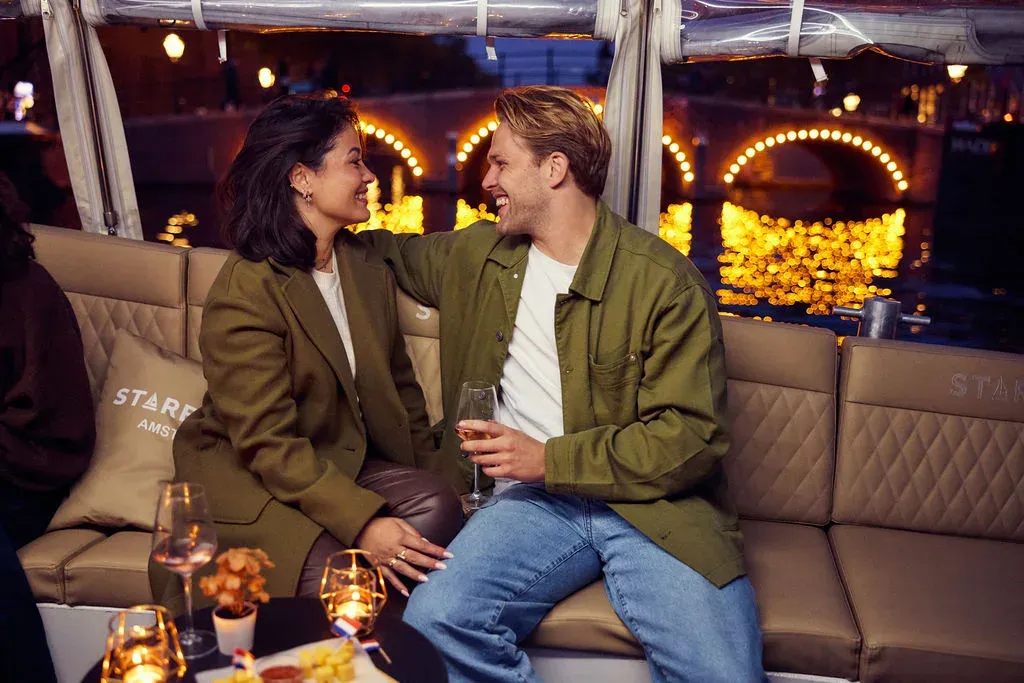 A couple sits close together with wine glasses in a covered Starboard boat, with a bridge outlined in lights behind them at night