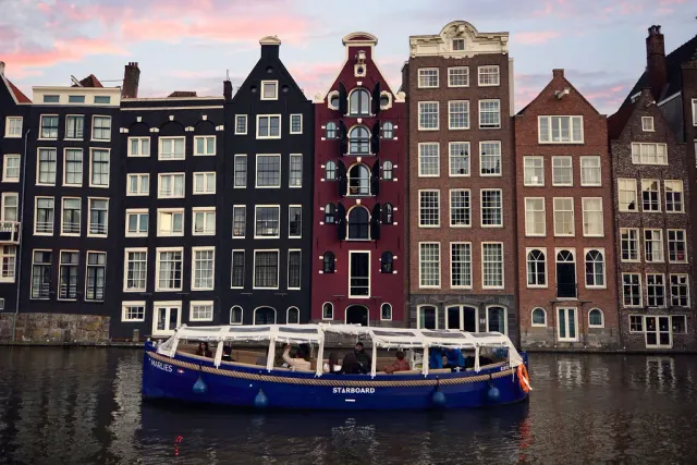 Covered blue Starboard canal boat with guests aboard in front of tall canal houses at dusk