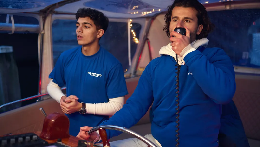 Two Starboard crew members in blue jackets at the helm at dusk, one speaking into a microphone