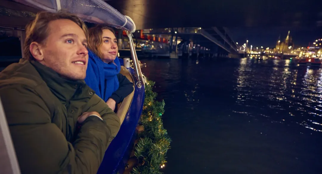 Guests in winter jackets look out over the water from the boat at night, with city lights in the distance
