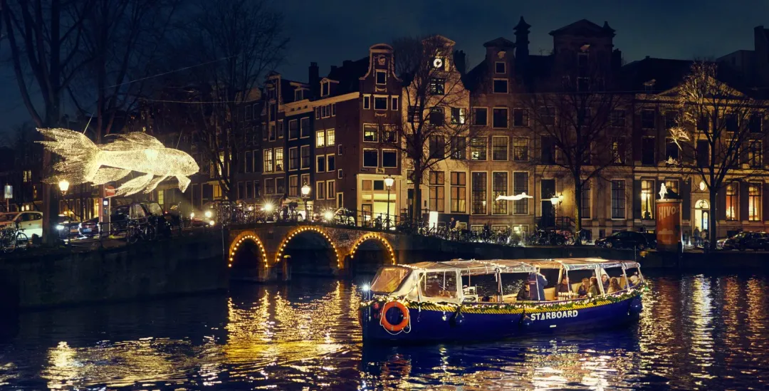 Starboard canal boat with a canopy on an Amsterdam canal at night, passing a glowing fish-shaped light sculpture and a lit arched bridge