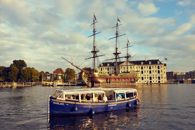 Covered blue Starboard canal boat with guests aboard on open water at sunset, in front of a historic tall ship