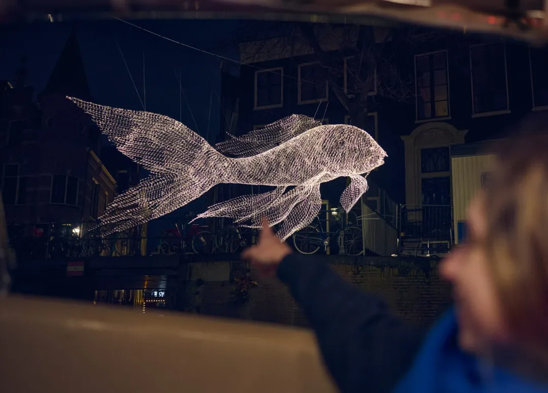 A wire-frame fish light sculpture glows above a canal at night, seen from a seat on a Starboard boat