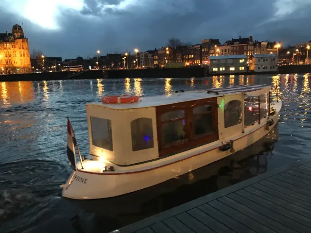 Cream-coloured canal boat Delphine moored at a wooden dock on the Amstel at dusk, with a lit cabin and city lights across the water
