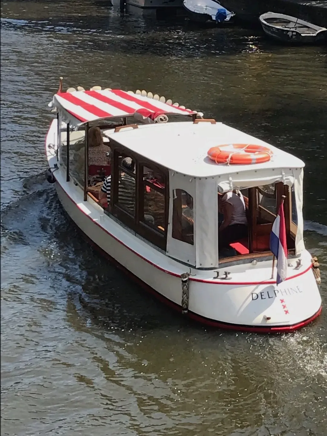 Small white canal boat Delphine with a red and white striped canopy moving along an Amsterdam canal
