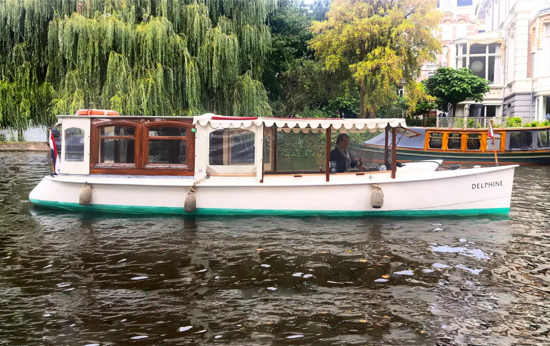 White canal salon boat Delphine with a wooden cabin and striped canopy cruising past willow trees on an Amsterdam canal
