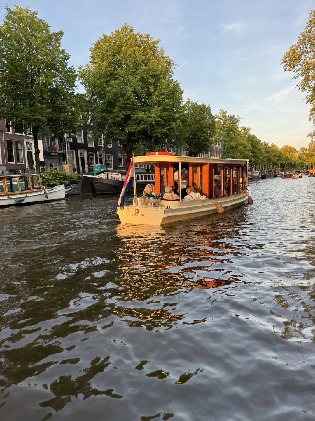Leaudine canal boat with a lit wooden cabin cruising along a tree-lined Amsterdam canal