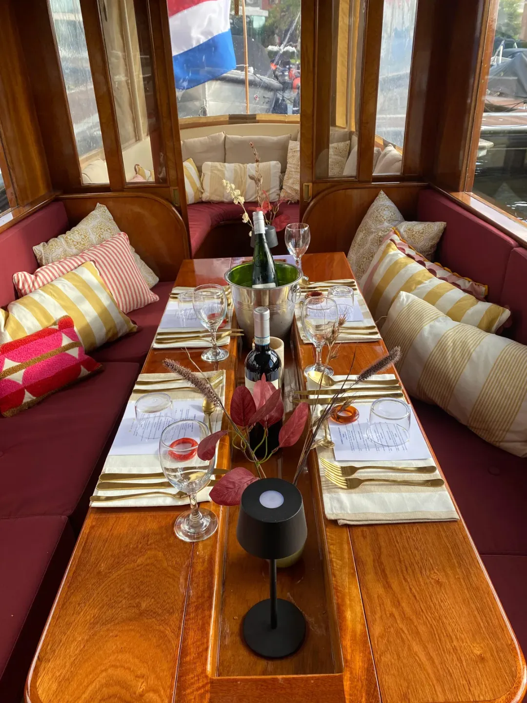 Long dinner table laid with glasses, wine and cutlery between red cushioned benches in a wooden canal boat salon