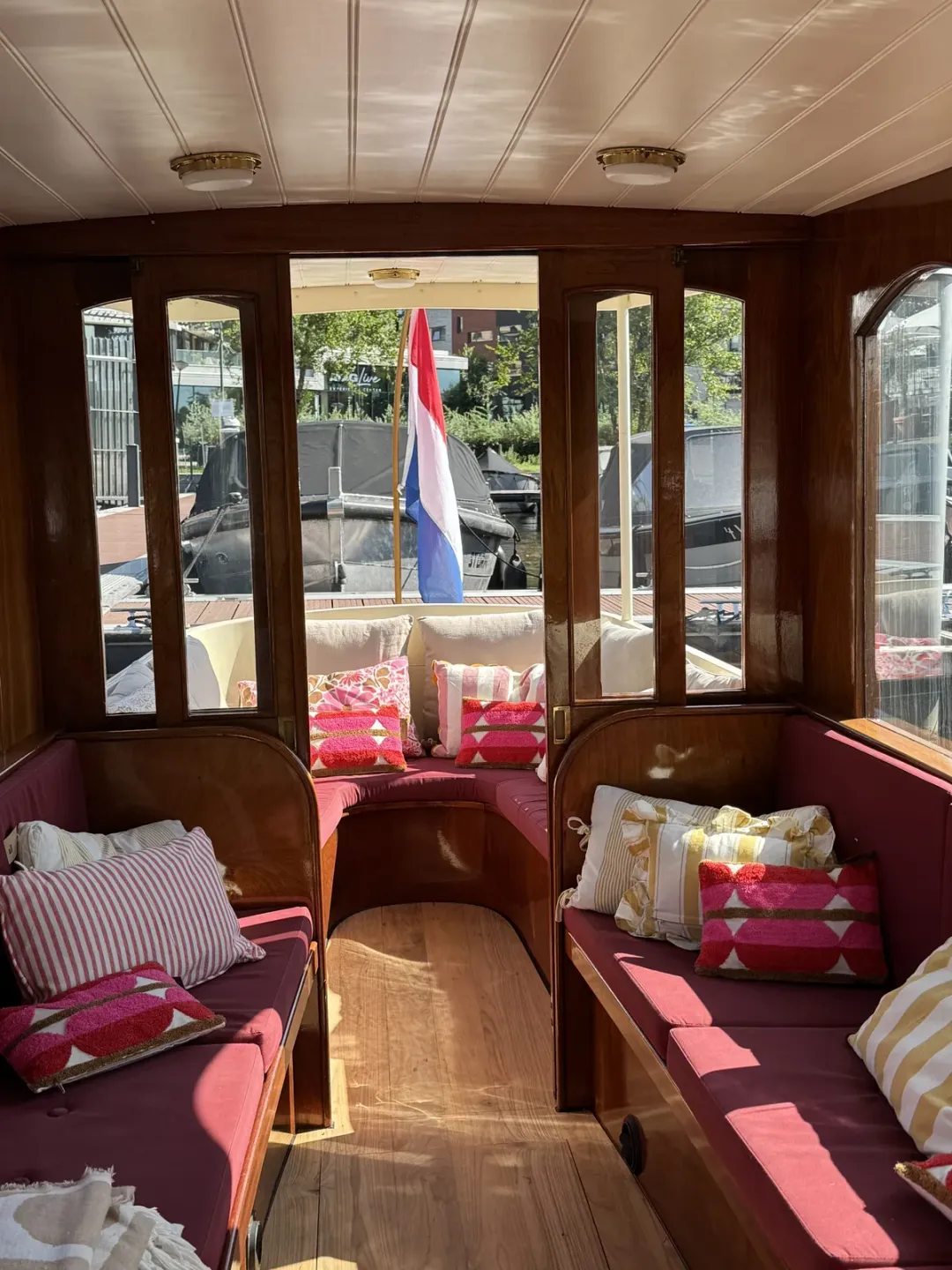 Wood-panelled boat salon with red cushions and striped pillows opening onto the aft deck, a Dutch flag outside
