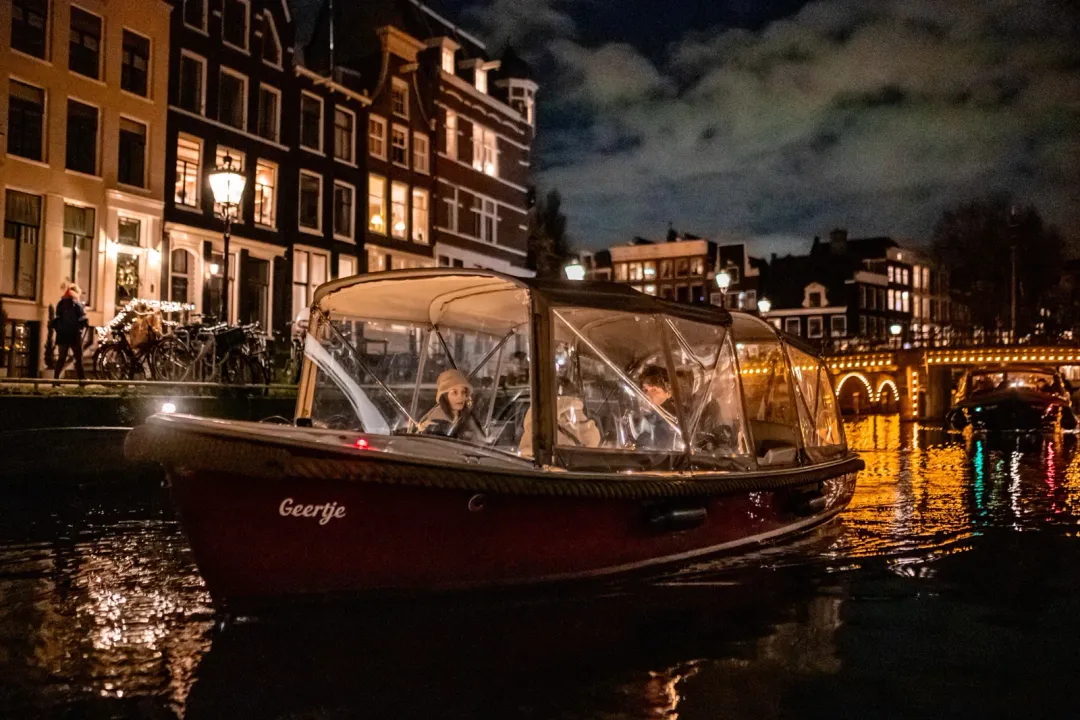 Red canal boat Geertje with enclosed canopy gliding past lit canal houses and an illuminated bridge at night in Amsterdam