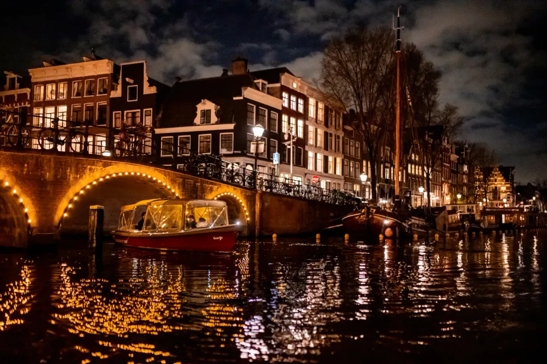 Red canal boat with glass canopy passing beneath a lit stone bridge at night in Amsterdam