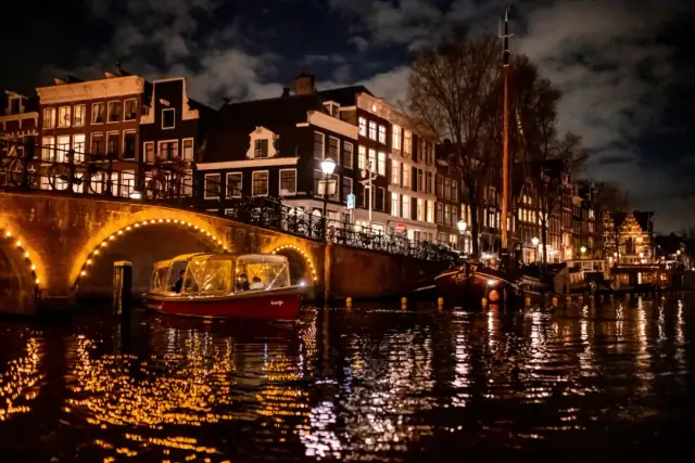 Red canal boat with glass canopy passing beneath a lit stone bridge at night in Amsterdam