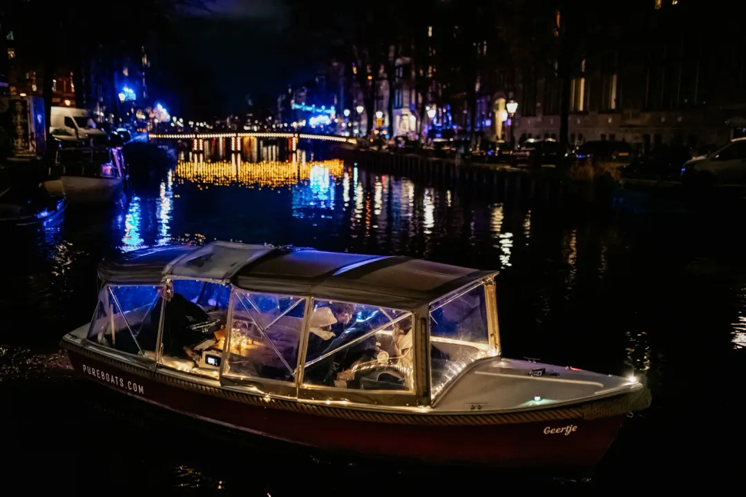 Red-hulled canal boat Geertje with a canopy glides through a dark Amsterdam canal beside a lit bridge
