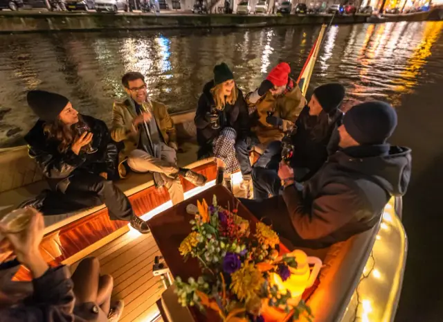 Friends in winter hats and coats sit around a flower-topped table on a boat at night on an Amsterdam canal