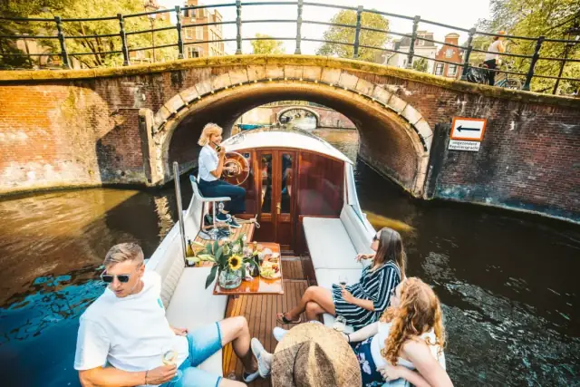 Guests sit in the open cockpit as the boat passes under a brick bridge, with the captain at the wheel behind them