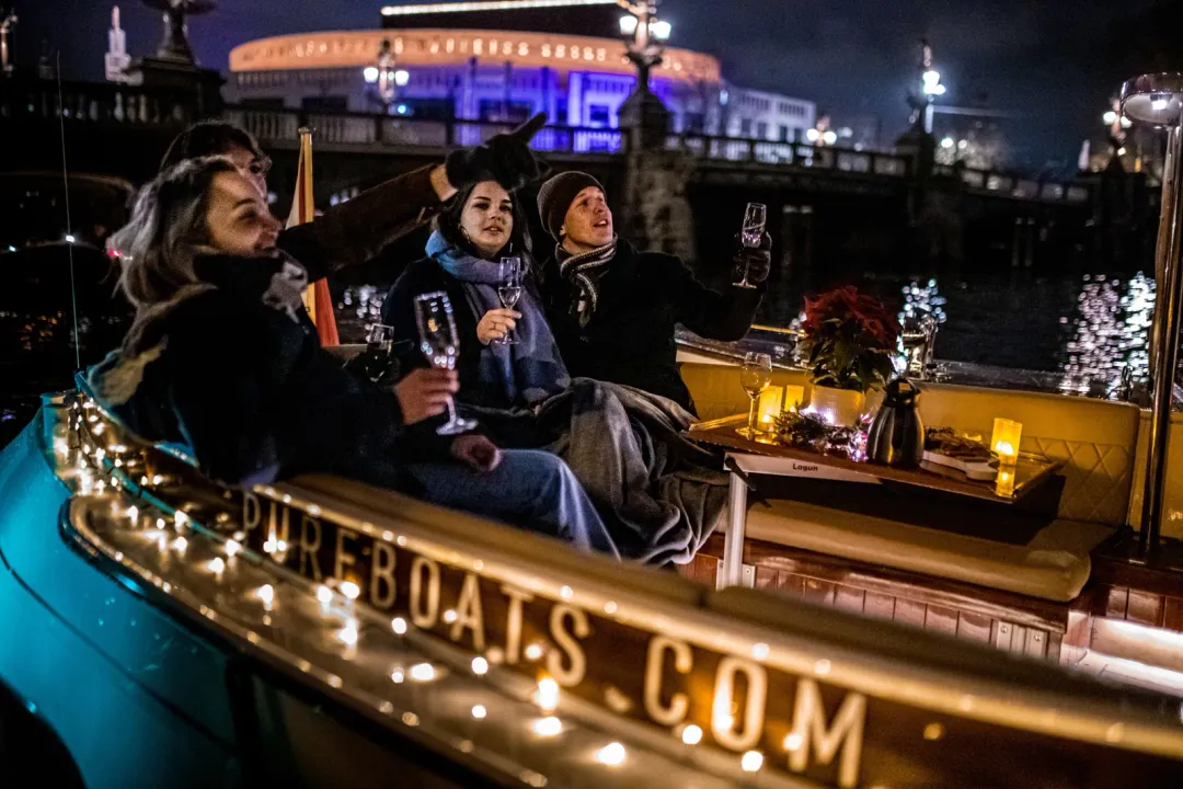 Guests in winter coats raise champagne glasses aboard a lit boat at night near an Amsterdam bridge