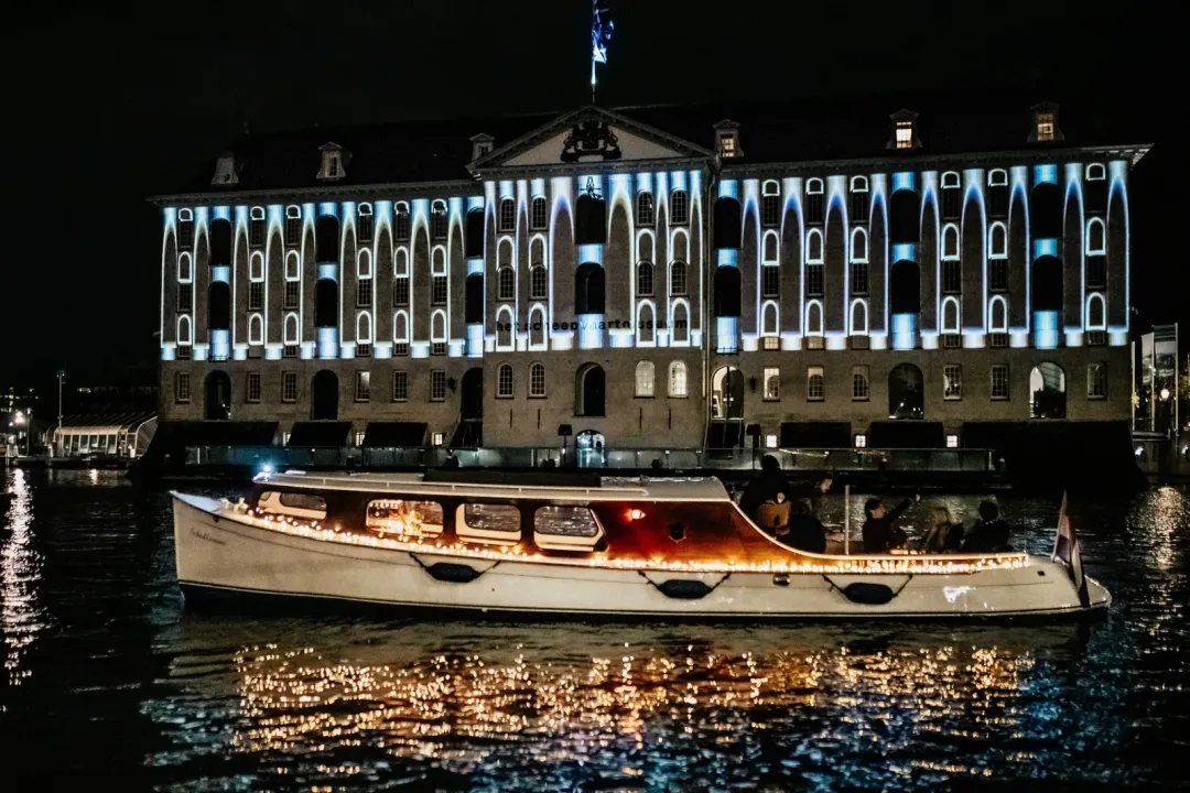 Schollevaar, a small cream saloon boat with lit windows, passes a canal building lit with blue light projections at night