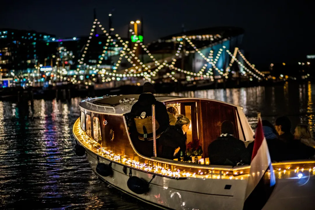 Schollevaar, a wooden saloon boat edged with fairy lights, carries passengers across Amsterdam's night-time canal