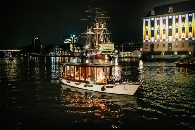Lucy, a lit-up wooden canal boat on dark water before a tall ship in Amsterdam
