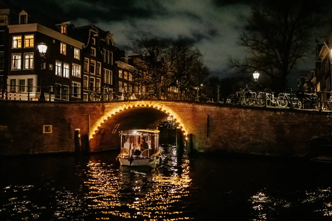 Small open boat Lucy passing beneath a lit brick bridge on an Amsterdam canal at night