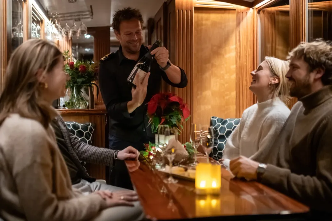 Smiling crew member in uniform shows a wine bottle to four seated guests at a wooden table