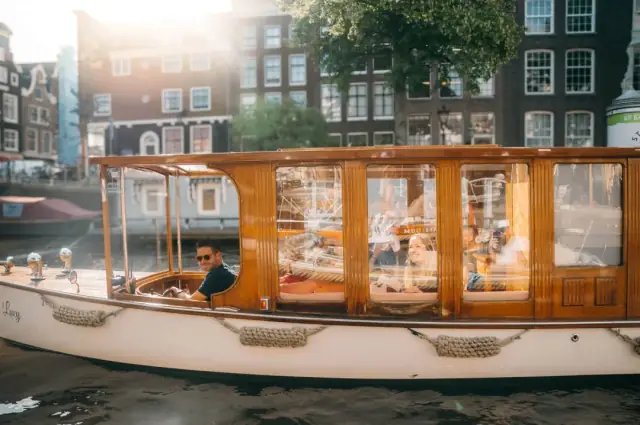 Lucy, a wooden canal boat with tall glass windows and the captain at the wheel, passes canal houses in sunlight