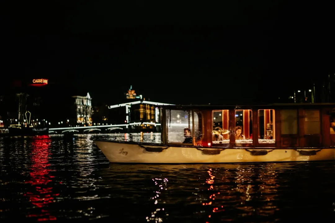 Wooden-windowed canal boat Lucy glides across dark Amsterdam water at night, passengers visible inside, Carré lit behind