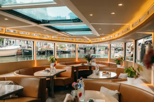 Boat interior with tan leather banquettes, round tables with flowers and glassware, skylight roof, canal visible through windows