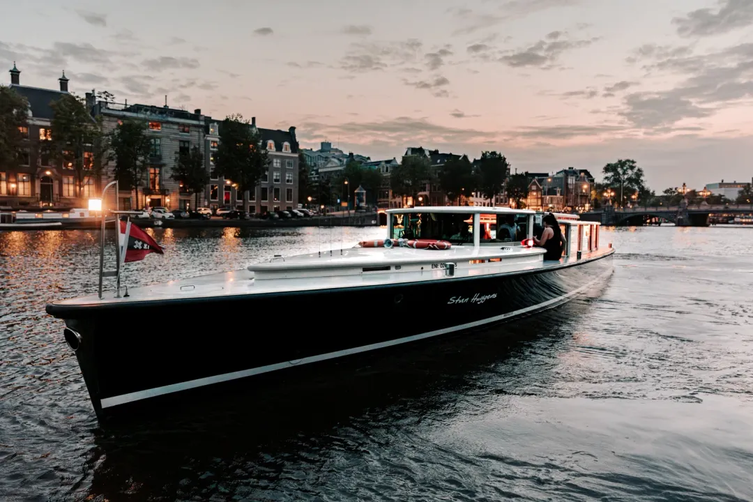 Stan Huygens, a dark-hulled canal boat, cruising the Amstel at dusk past lit canal houses