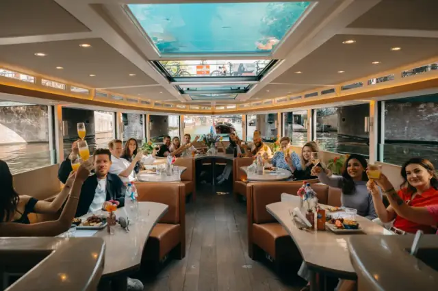 Guests raise glasses in a toast at tables inside a glass-roofed canal boat