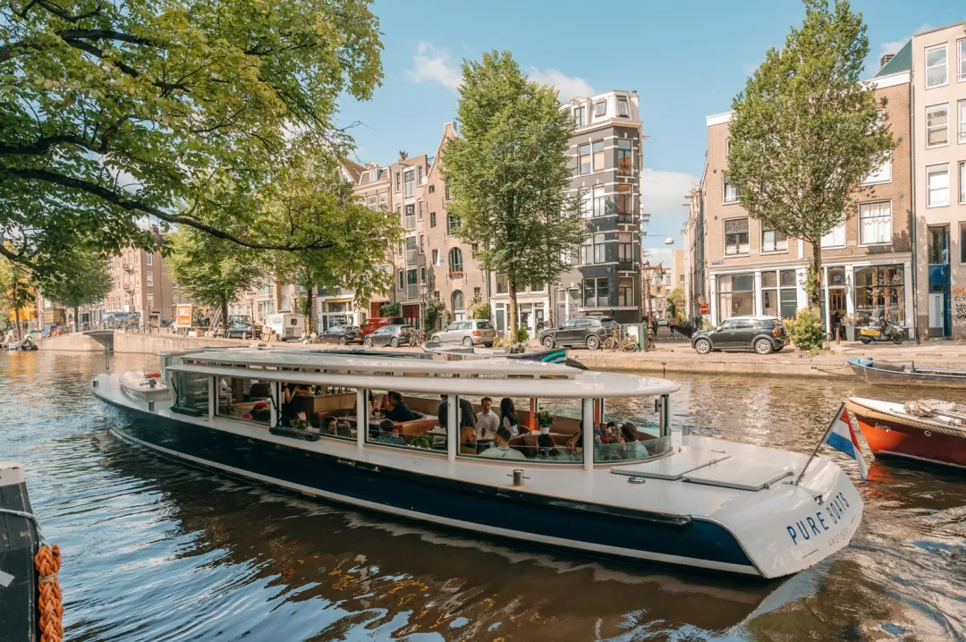 Navy and white glass-roofed canal boat with seated passengers moving along an Amsterdam canal lined with trees and tall brick houses