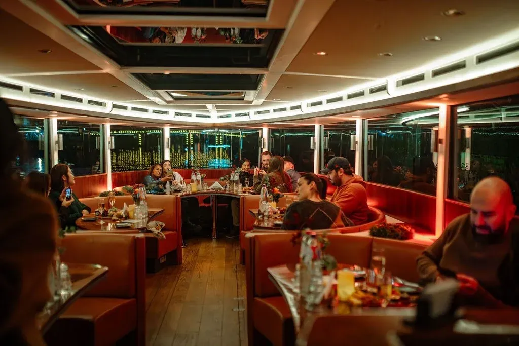Guests seated in orange leather booths at candlelit tables inside a glass-roofed boat after dark