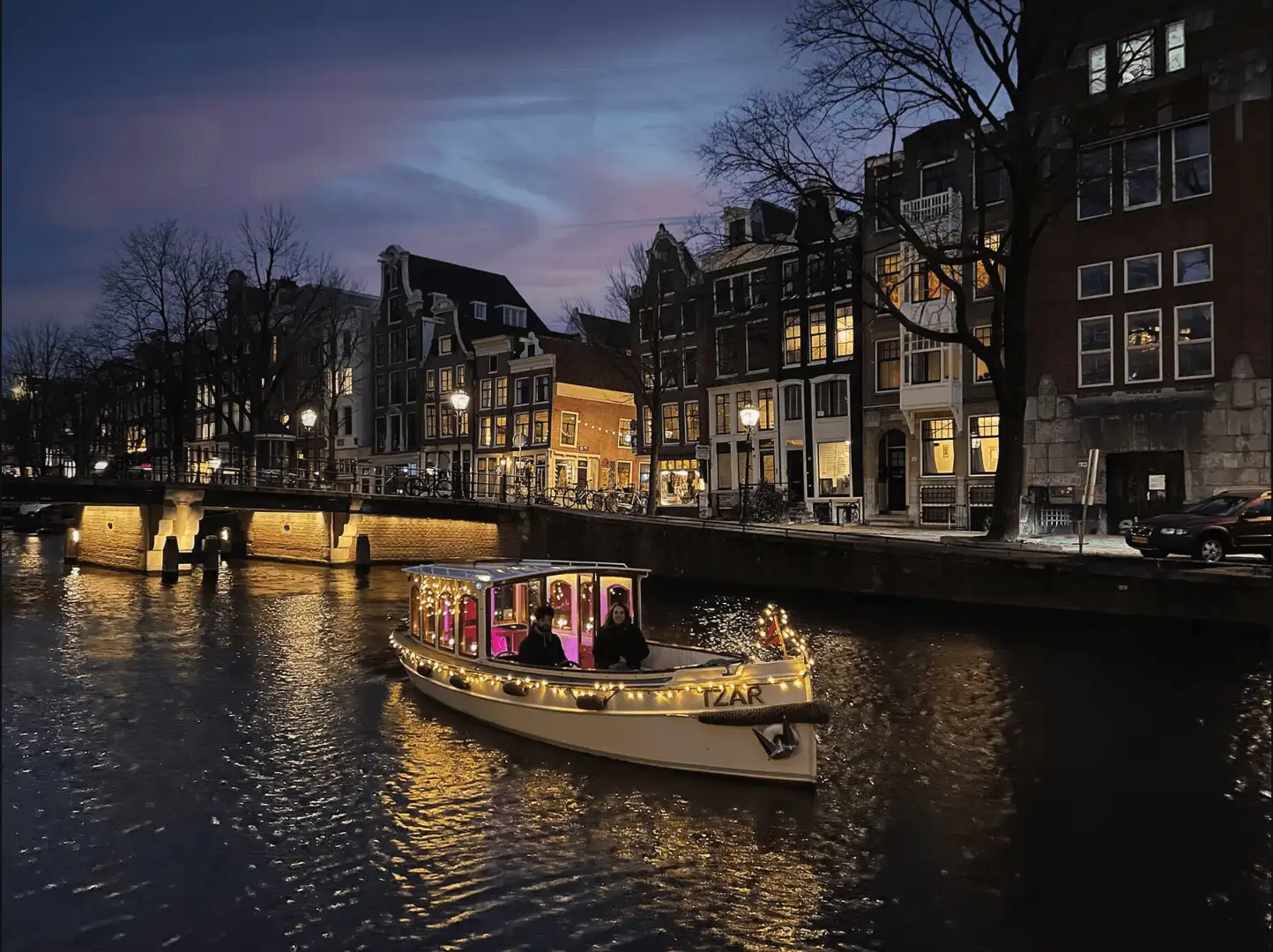 The saloon boat Tzar lit with string lights on a canal at blue hour, canal-house windows lit behind it