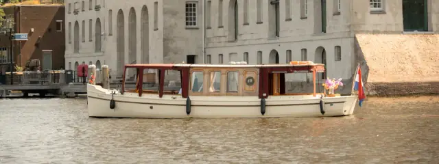 Cream-hulled 1910 salon boat Dauphine with a dark red canopy moored on an Amsterdam canal, Dutch flag at the stern