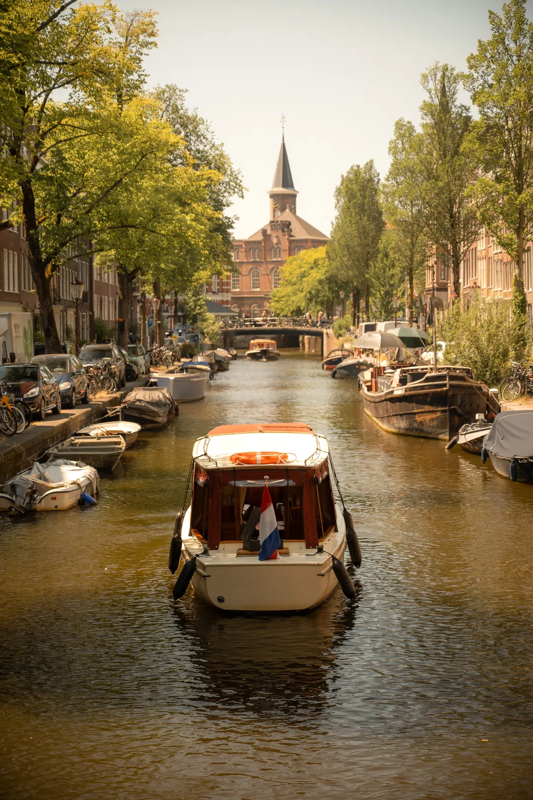 Salon boat with a Dutch flag sailing down a tree-lined Amsterdam canal toward a church tower