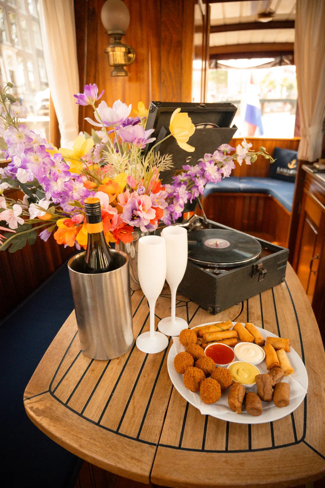 Wooden table in the salon with a wine cooler, white glasses, a vintage record player, flowers and a plate of Dutch snacks