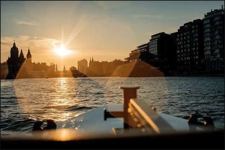 The sun setting over open water near Amsterdam Centraal, the twin towers and dome of the Basilica of St Nicholas in silhouette, seen from a small boat