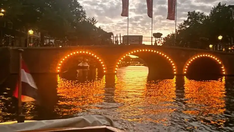 A three-arched Amsterdam canal bridge at sunset with small lights around each arch, reflected in the water