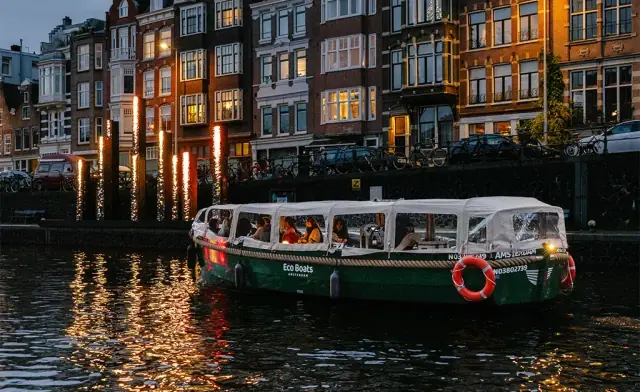 Green Eco Boats canal boat with a covered cabin and passengers aboard, on an Amsterdam canal at dusk with lit houses behind