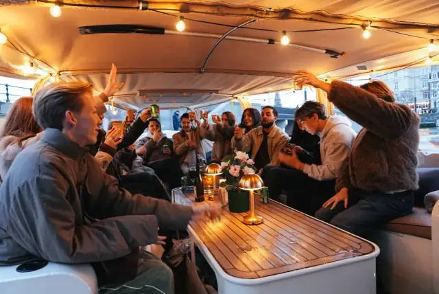 Group of guests raise their drinks inside a covered boat under string lights, with a table lamp and flowers on the table