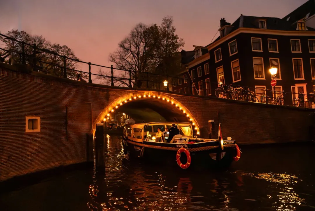 Eco Boats canal boat with a lit cabin and orange lifebuoys passing under a brick bridge arch outlined with warm lights at night