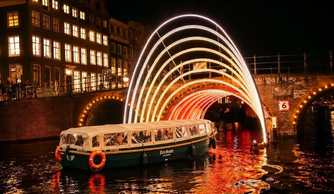 Eco Boats canal boat with passengers passing beneath a glowing arch of white and orange light on an Amsterdam canal at night