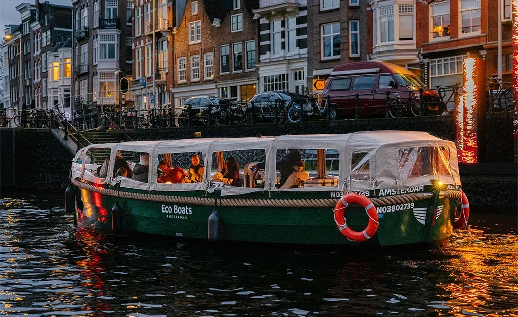 Green Eco Boats canal boat with a covered cabin, passengers aboard and a lifebuoy on the side, on an Amsterdam canal in daylight
