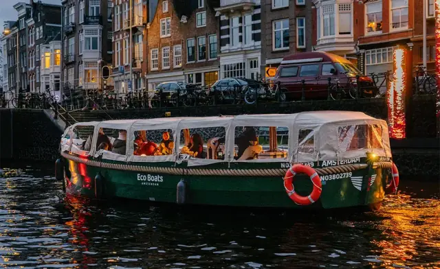 Green Eco Boats canal boat with a covered cabin, passengers aboard and a lifebuoy on the side, on an Amsterdam canal in daylight