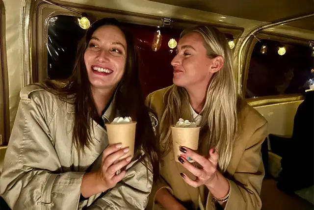 Two smiling women in trench coats hold paper cups of hot drinks topped with cream inside a boat cabin at night