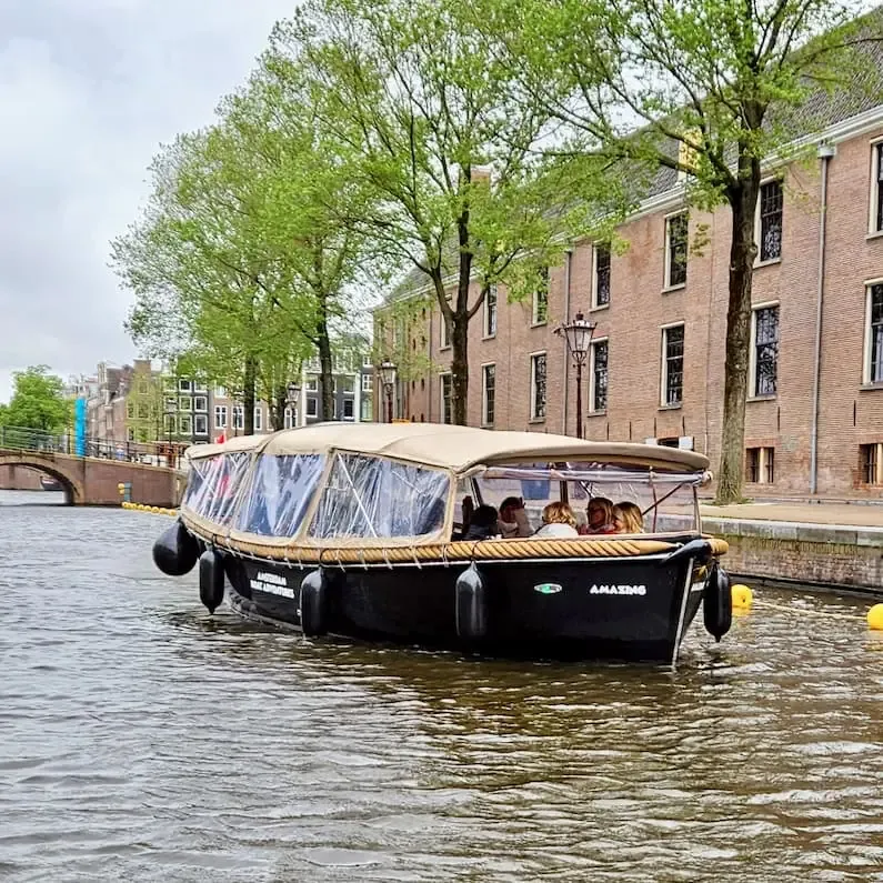 Open canal boat with the clear rain cover fitted, guests seated inside, passing a brick building and trees in Amsterdam