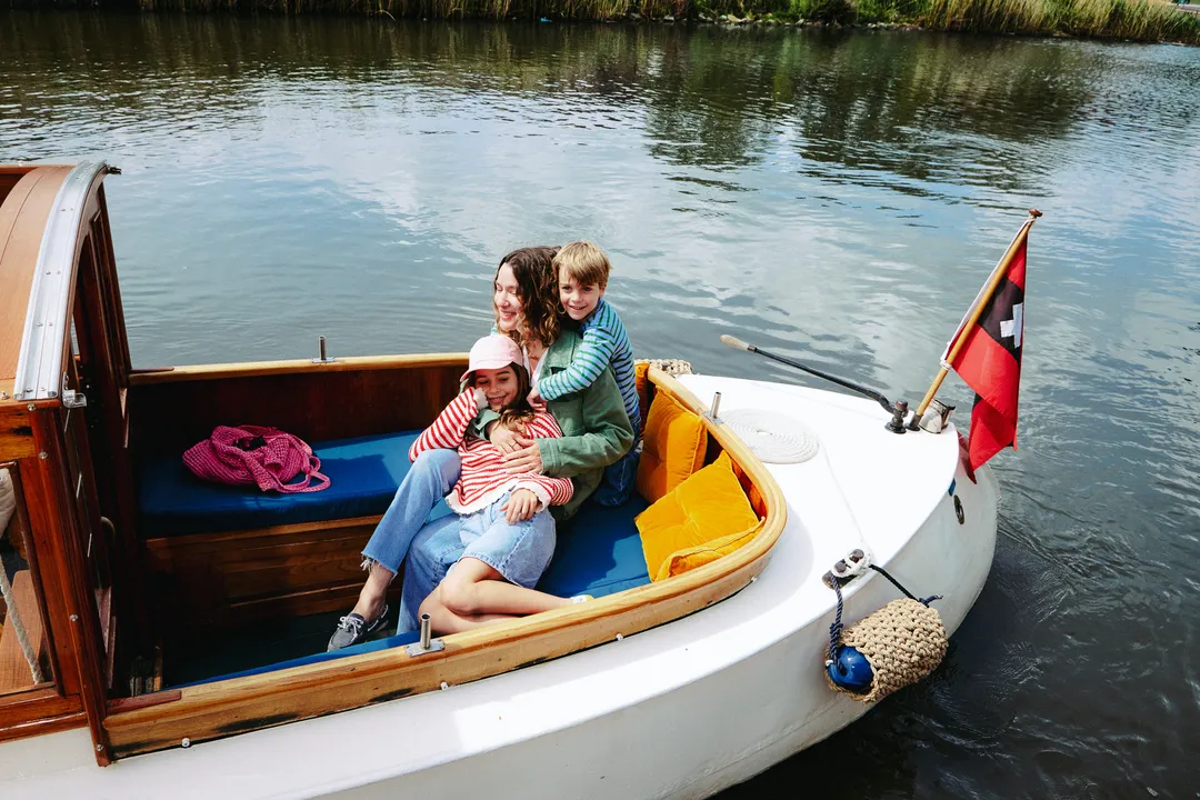 Woman and two children sit close together on blue cushions in the open cockpit of Diana, a white wooden boat