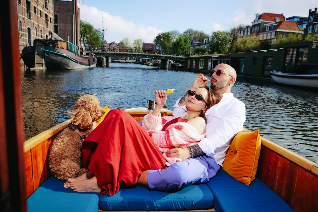 Couple in sunglasses reclining on blue cushions with a curly brown dog on an Amsterdam canal