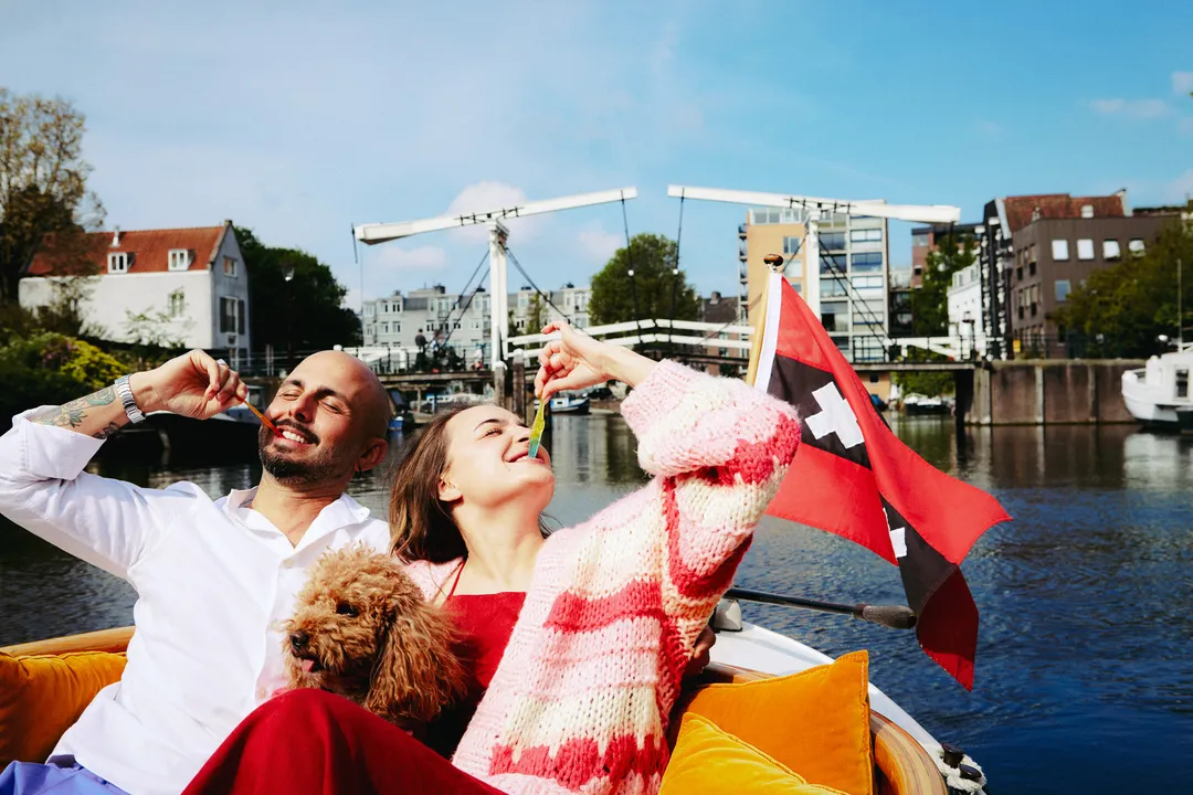 Laughing couple with a small brown dog lounge on orange cushions, Amsterdam flag and drawbridge behind
